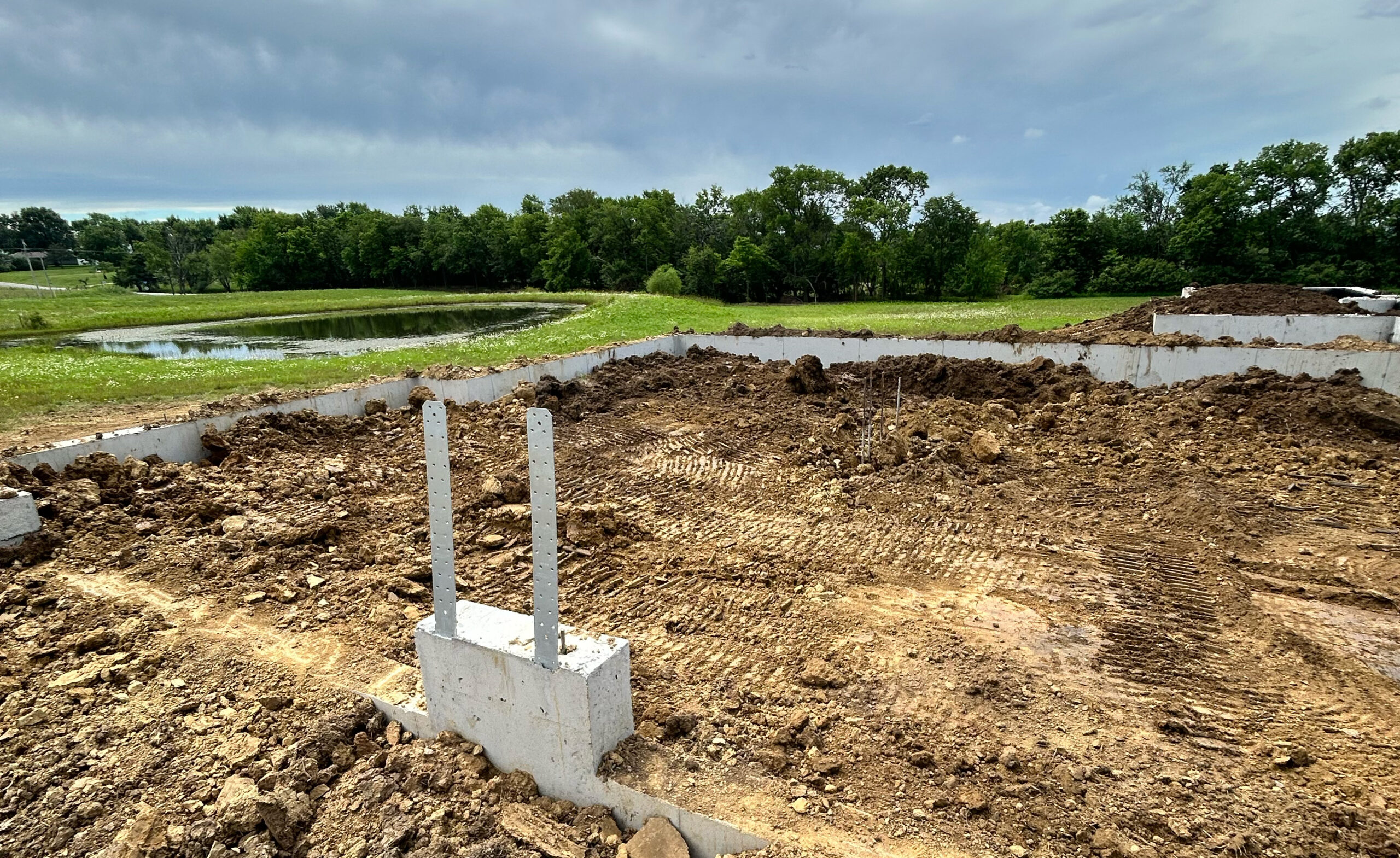 Concrete foundation in dirt by a pond and tree line.