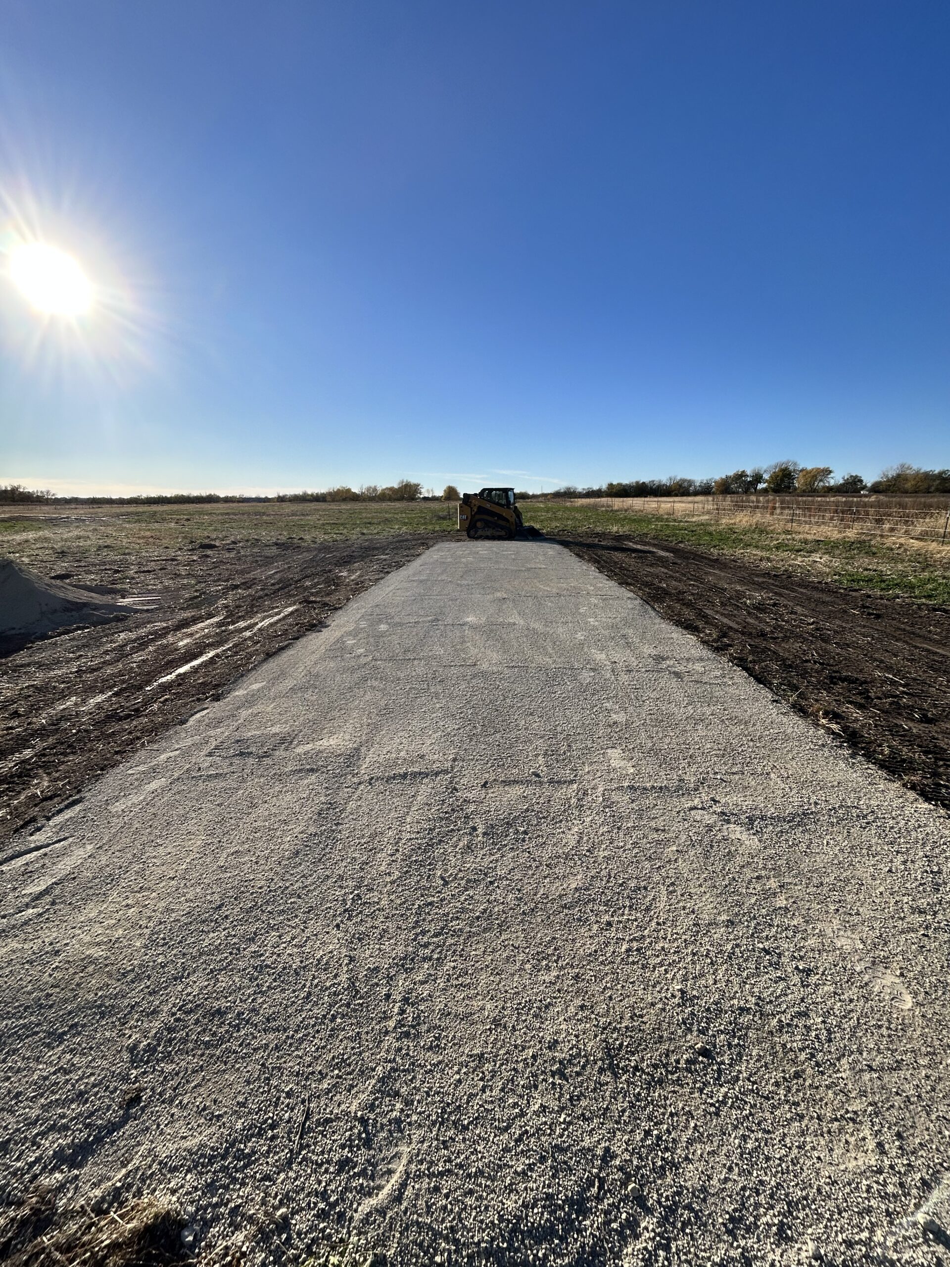 Gravel driveway with skid steer under bright sun and blue sky.